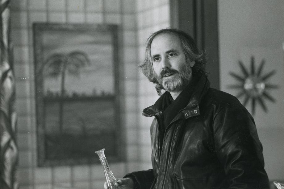 bearded man in a leather jacket holding a glass bud vase