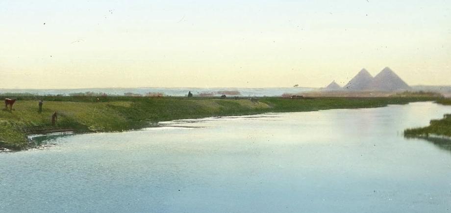 A view along the length of a wide river with the peaks of pyramids in the distance.