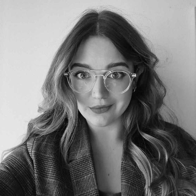 Black and white photo of a white woman with long hair, round glasses, and a checkered blazer.