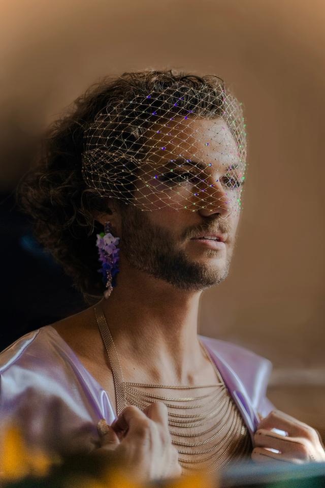 Close-up of j. Oceano — with chin-length dark curly hair and light facial hair, wearing a sparkling constellation veil draped across their face. They gaze slightly off to the side with a calm, contemplative expression, wearing lilac floral statement earring, layered gold chains, and a shimmering purple satin garment held lightly at the collar. The background is softly blurred in warm beige tones, with gentle lighting emphasizing the veil's kaleidoscopic reflections.