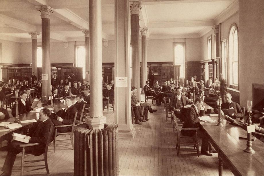 a large room with long tables and men seated around them reading