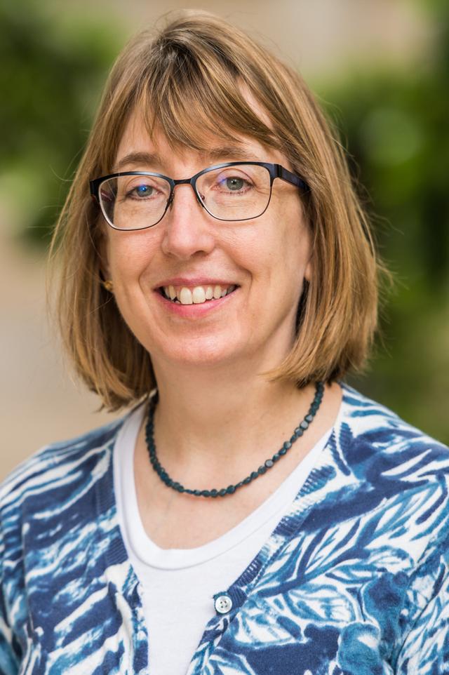 Photo of Chris Powell, a middle-aged woman with glasses and chin-length light brown hair, wearing a white shirt, a blue necklace, and a blue and white cardigan.