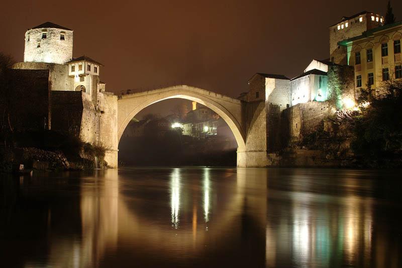 Arched bridge connecting concrete buildings over a river with a reflection in the water of the city in the distance.