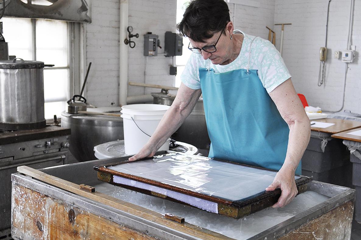 Woman pulling a large wooden frame up out of a water bath, making paper.
