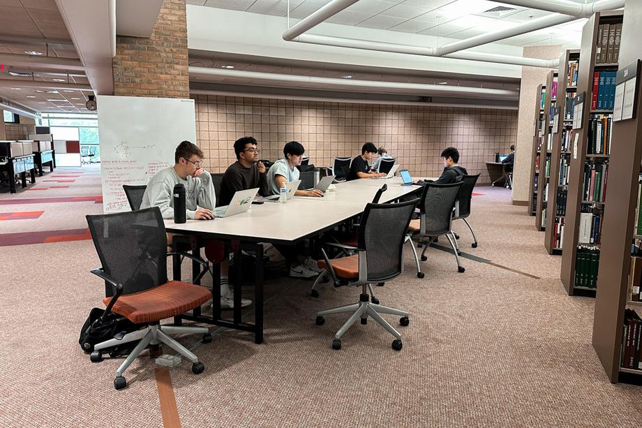 Students working at a long rectangular table next to book stacks with a whiteboard behind them and windows in the distance.