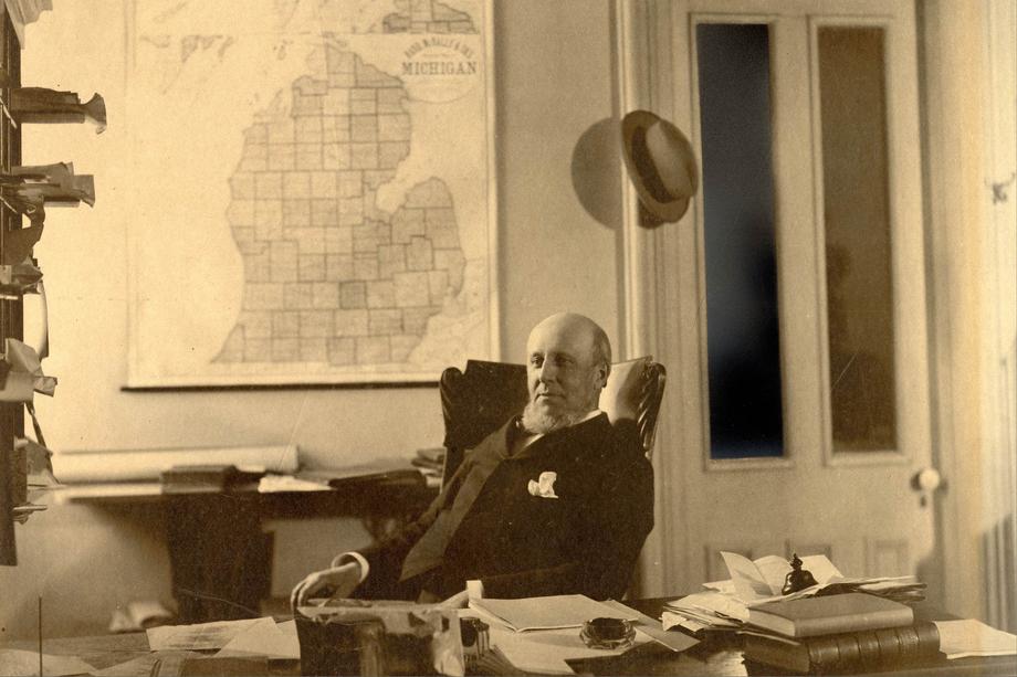 man seated at a desk with a map of Michigan on the wall behind him