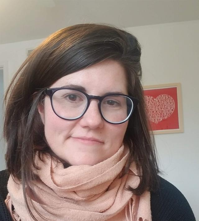 Photograph of Heidi Burkhardt, a middle-aged, white woman with dark brown shoulder length hair and glasses, smiling with her head tilted, and wearing a pink scarf and black sweater.