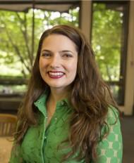 Photo of Maura Seale, smiling white woman with long curly brown hair, wearing a green dress, and sitting in front of a window that looks onto trees.