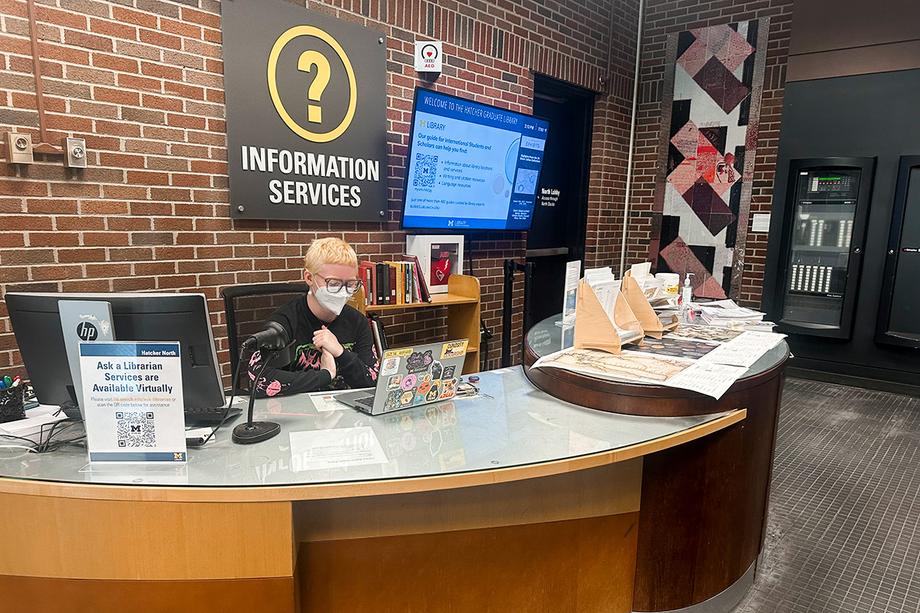 A masked library employee staffing the information desk.