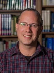 Photograph of Aaron Elkiss, a white person wearing glasses and a plaid button-down shirt, standing in front of a bookcase.