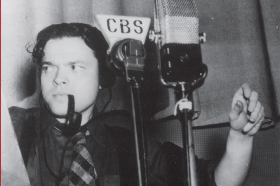 Actor/Director Orson Welles with a pipe in his mouth in front two microphones, one labeled CBS.