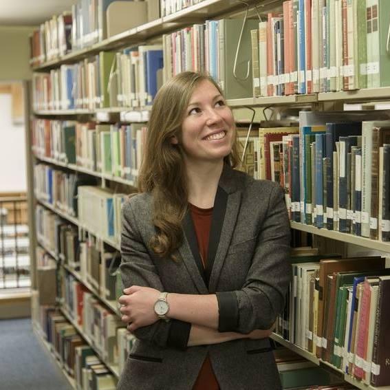 Woman in grey blazer stands in front of bookshelf, smiling