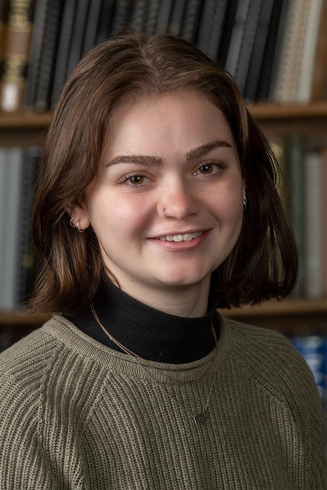 A white woman with shoulder-length brown hair, smiling in front of a shelf of books