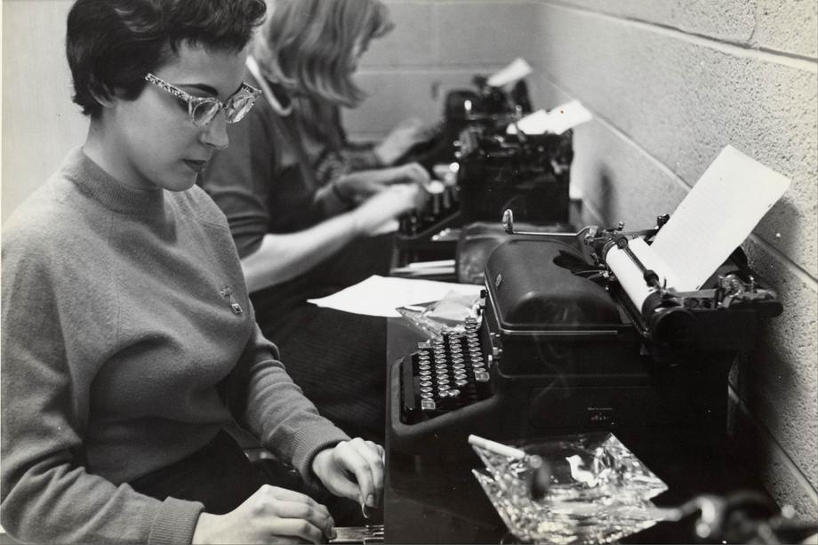 woman seated at a typewriter which sits beside an ashtray