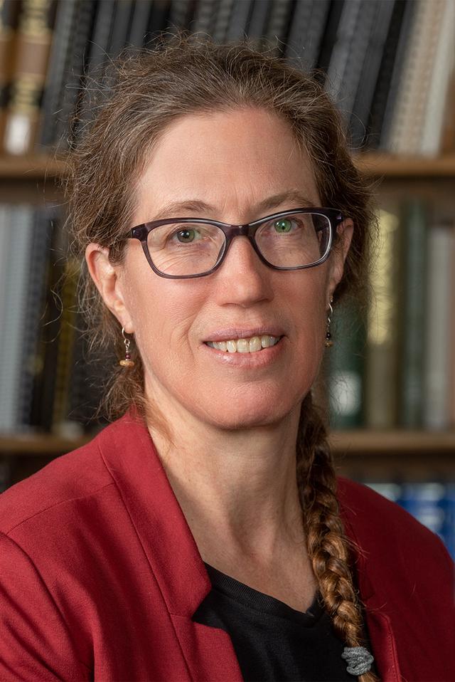 Photograph of Kat Hagedorn, a white woman with braided brown hair wearing clear glasses and a red blazer, in front of a bookshelf.