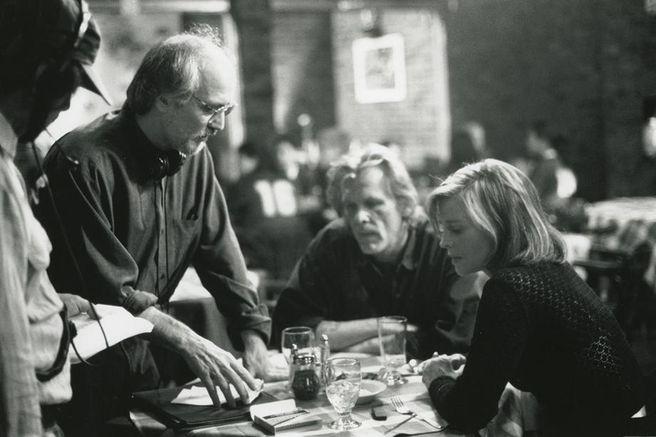 Man in glasses standing and talking to a man and woman seated at a table