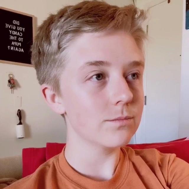 A photo of Annie cropped to show them from the neck up.  They have blond hair and a short, &quot;boyish&quot; haircut and are wearing an orange crewneck shirt.  They are looking offscreen to the right.