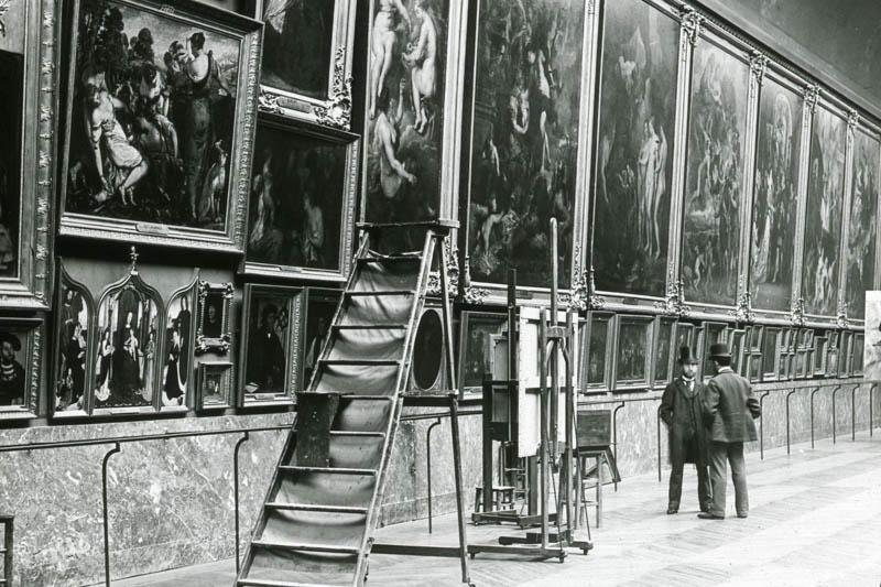 Black and white photo of a gallery high ceilings, paintings hanging on the wall, and people looking at them.