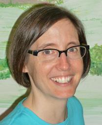 Photograph of Beth Snyder, a white woman with short brown hair wearing clear glasses and a turquoise top, in front of painted mural.