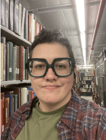 A picture of Shannon Moreno. A person with short dark hair, large square green glasses, wearing an olive green shirt with burgundy flannel, and standing in front of books. 