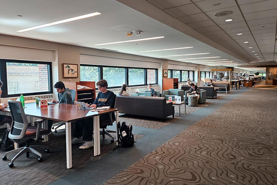 Students studying at rectangular tables in comfy chairs alongside windows.