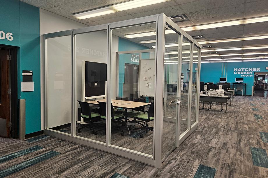 Glassed in space along a wall with a table, five chairs, and wall-mounted screen. Computers and entrance to Hatcher South are in the background.