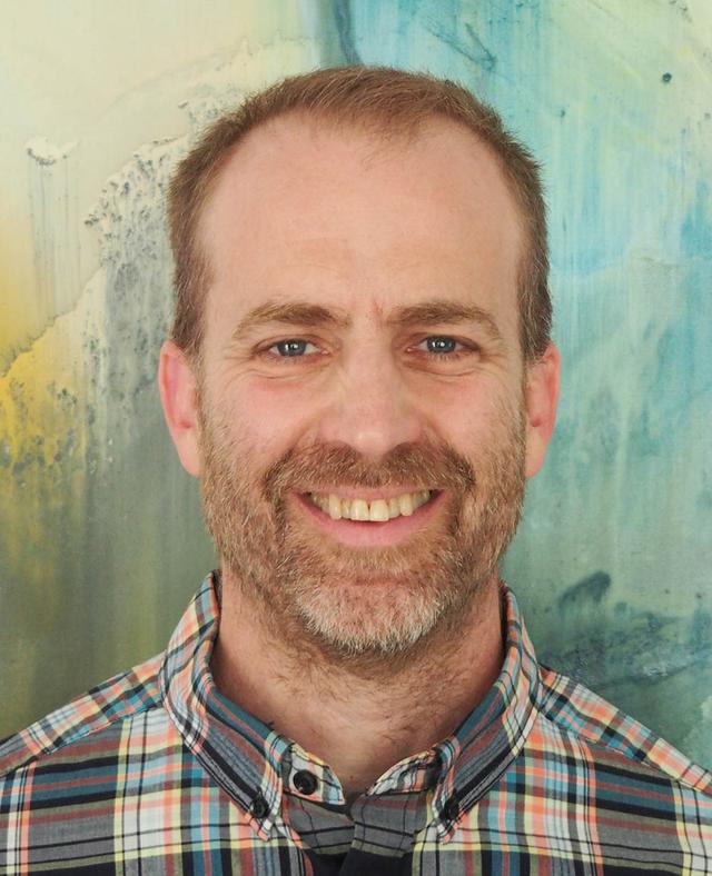 A smiling, white, older male with short blond hair and beard, wearing a chequered shirt, standing against a multicolor abstract background.