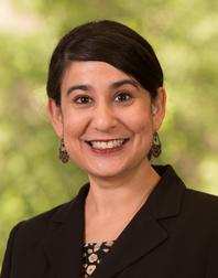 Photograph of Gurpreet Kaur Rana, a woman of South Asian heritage standing in front of light green foliage, wearing a black blazer and with a smile.