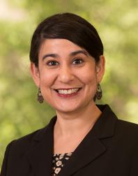 Photograph of Gurpreet Kaur Rana, a woman of South Asian heritage standing in front of light green foliage, wearing a black blazer and with a smile. 