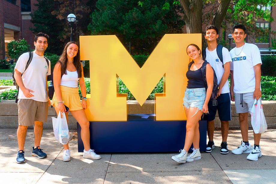 Students posing next to a large block M with trees and the Hatcher Library in the background