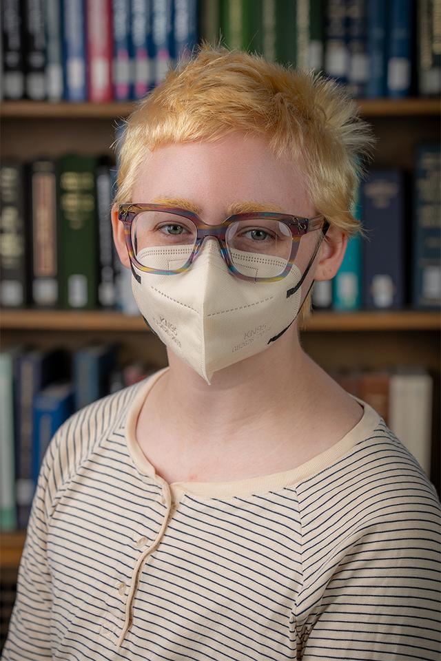 A white person with cropped bleached hair, bleached eyebrows, large glasses, and wearing a face mask