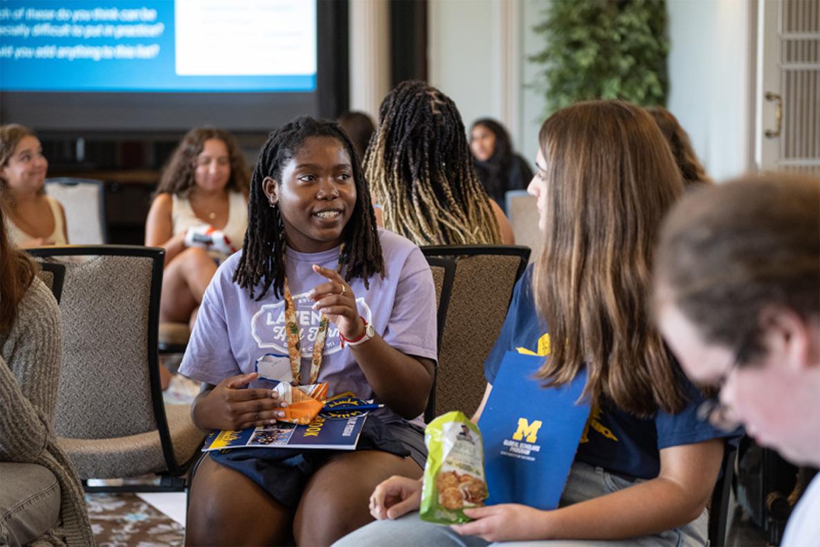 A group of students sit and talk in a classroom setting, holding University of Michigan materials and snacks, with a presentation screen visible in the background.