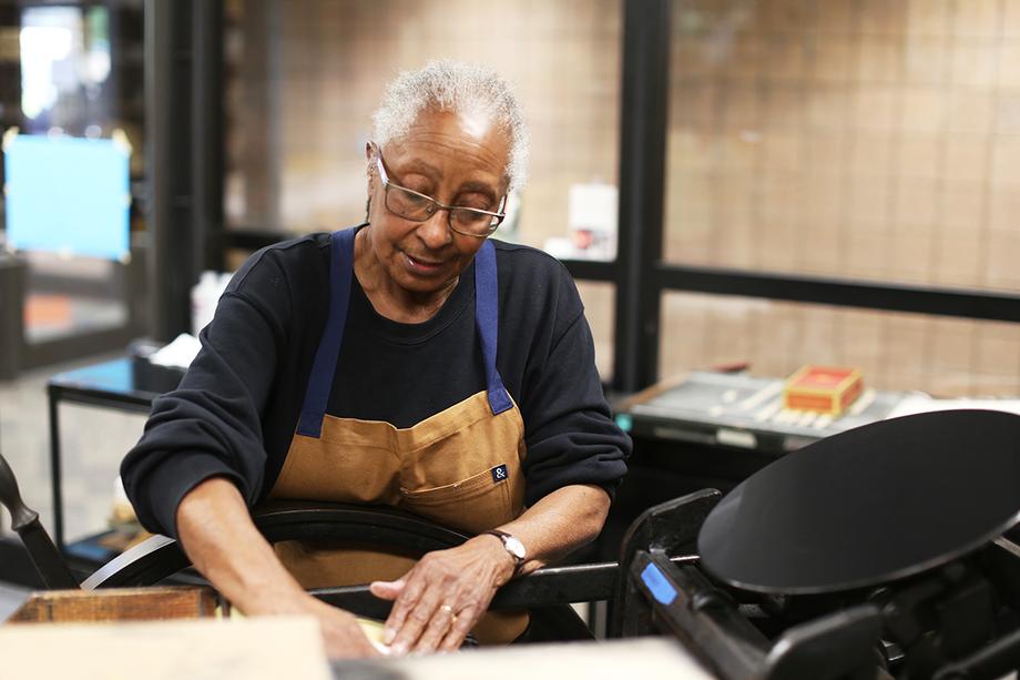 Woman wearing a leather apron and working at a printing press.