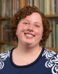 Photograph of Leigh Billings, a white woman with curly hair wearing a blue and white shirt and standing in front of a bookshelf