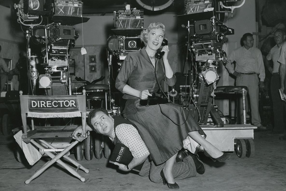 A man on his knees next to a director's chair, and a woman using his back as a chair while she talks on a telephone.