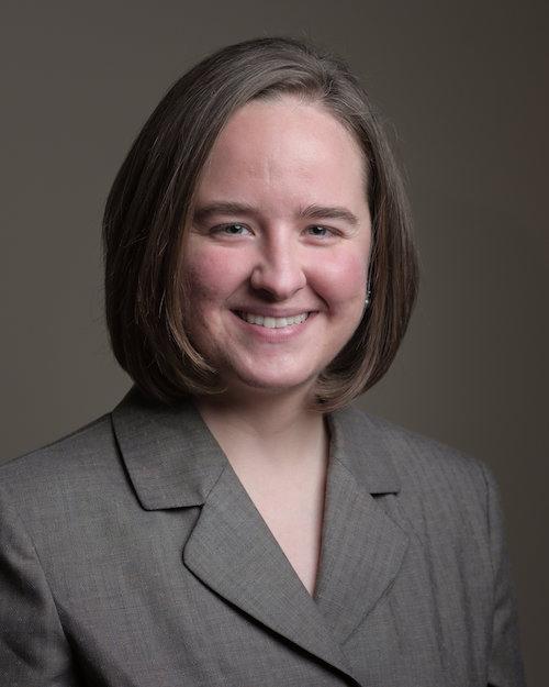 Photo of Sara Samuel. She is a thirty-something white woman with medium length light brown hair, wearing a light brown suit top and is smiling.