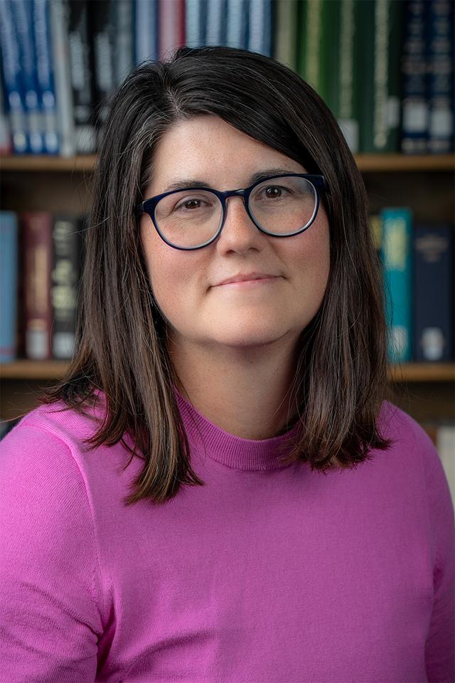 Photograph of Heidi Burkhardt, a middle-aged, white woman with dark brown shoulder length hair and glasses, smiling with her head tilted, and wearing a bright pink sweater.