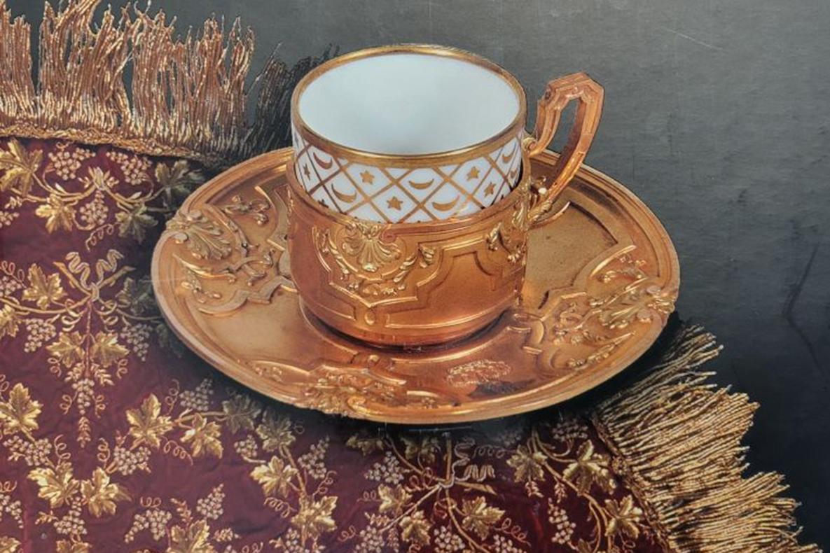 Ornate gold coffee cup on a matching gold saucer, sitting on a red and gold textile decorated with grape vines.