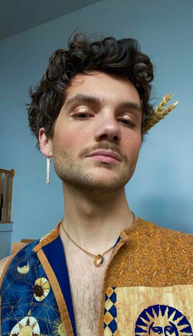 Close-up of j. Oceano — with short, dark curly hair and light facial hair, wearing shimmery gold eye makeup. They look slightly down at the camera with a neutral expression, wearing one long gold earring, a thin gold chain necklace with a small ring pendant, and a patterned vest in blue and gold with sun-and-moon motifs. A sprig of dried wheat is tucked behind their head on the right; pale blue wall and a wooden stair rail in the background.