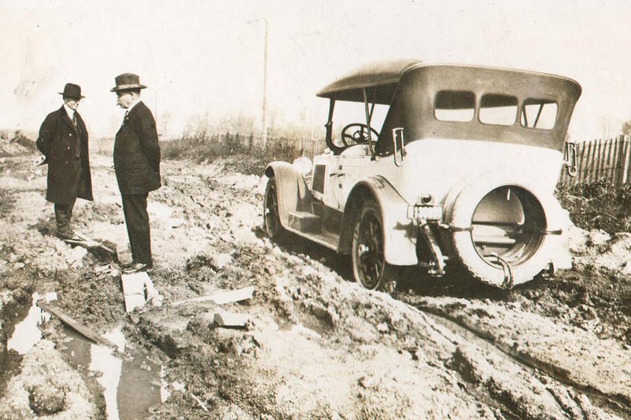 Two men in suits and hats standing in a muddy road next to a car