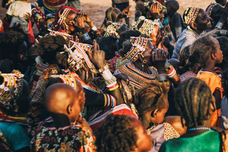 A group of people sitting on the ground in a town in Kenya, wearing vibrant clothing and beads.