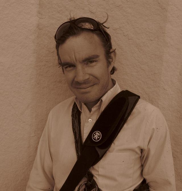 Photograph of Ben Howell, white male with short hair and a light button down shirt, standing outside against a light rock wall. 