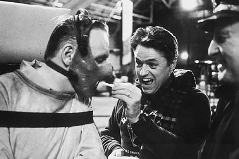 man extending a cigarette to another man in a mask and a straight jacket
