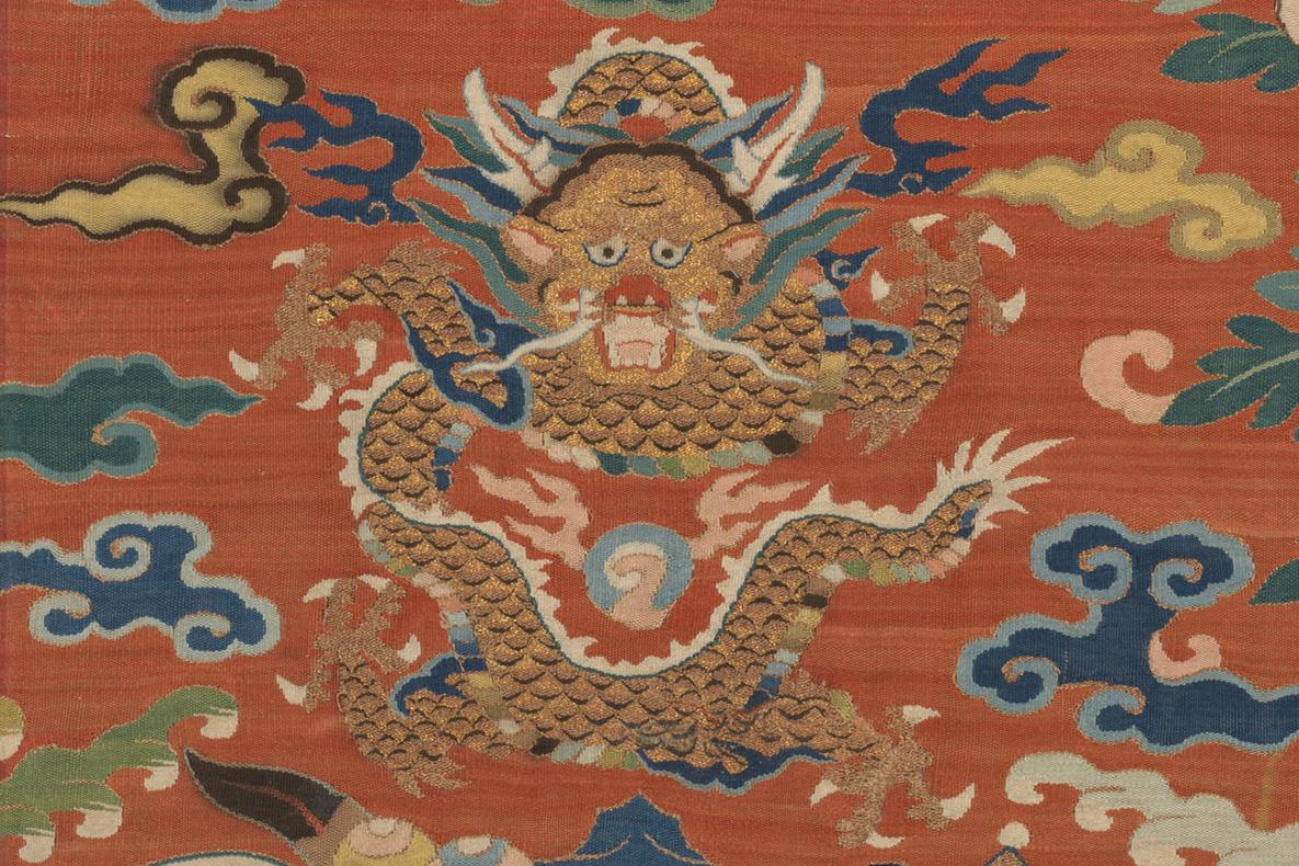 Part of a colorful silk tapestry, lavishly embellished with gold thread, depicting a coiled dragon staring directly at the viewer. 