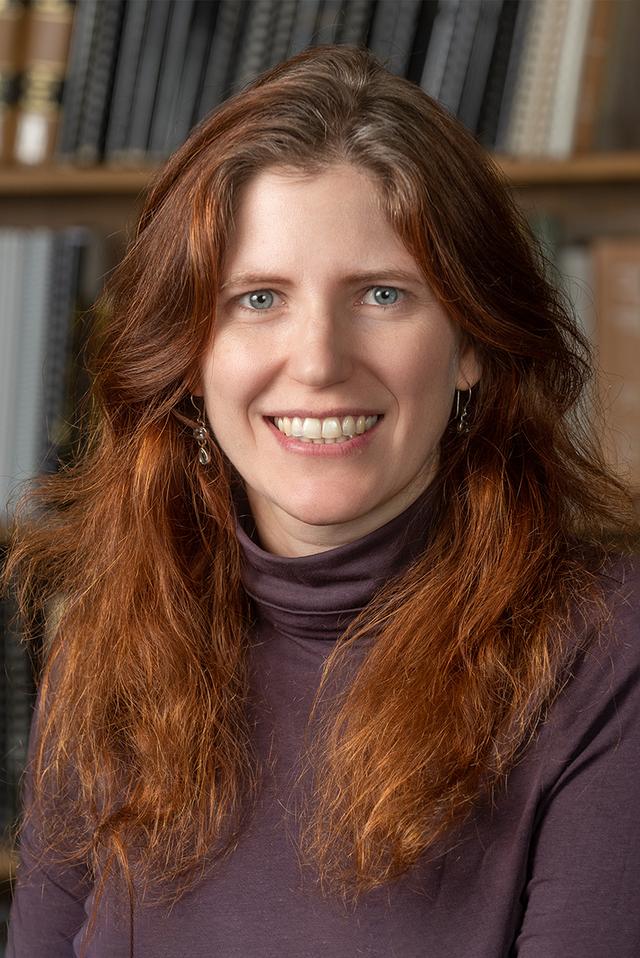 Photograph of Juli McLoone, a white woman with red hair, dark blonde at the roots, and blue eyes, wearing purple.