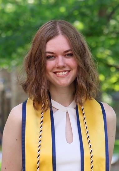 A woman with medium-length, wavy brown hair, smiling. She is wearing a white dress, a yellow graduation stole, and a blue, white, and yellow graduation cord, and she is standing outside in front of trees.