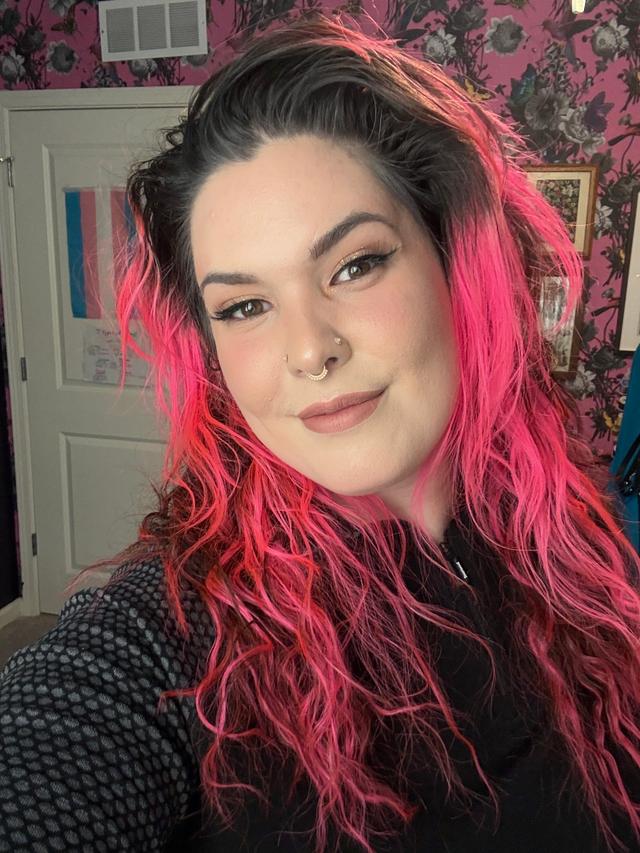 Smiling woman with light colored skin, peach colored lips, dark hair with bright pink highlights, and wearing a black shirt.