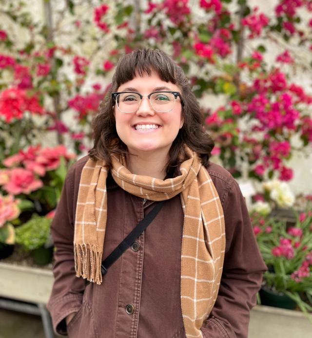 White woman with shoulder length brown hair and glasses wear a brown coat and tan scarf, standing against a background of red flowers.nt background. 