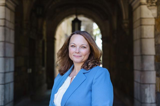The picture is located near an archway to the Law Quadrangle at the University of Michigan's Ann Arbor campus. I am a white female with shoulder-length light brown hair, and I have blue eyes. I am wearing a light blue suit jacket with a white shirt underneath.