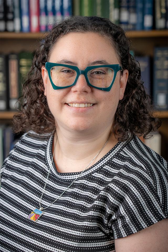 Brunette woman with teal glasses smiling. Shown shoulders up in a white striped dress with books in the background.